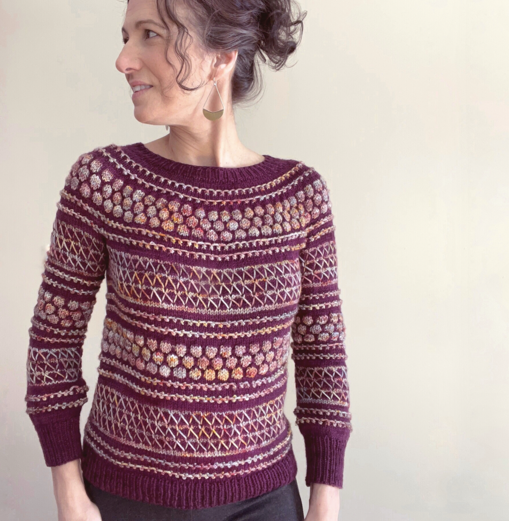 A light-skinned woman wears a purple and multicolored handknit sweater.