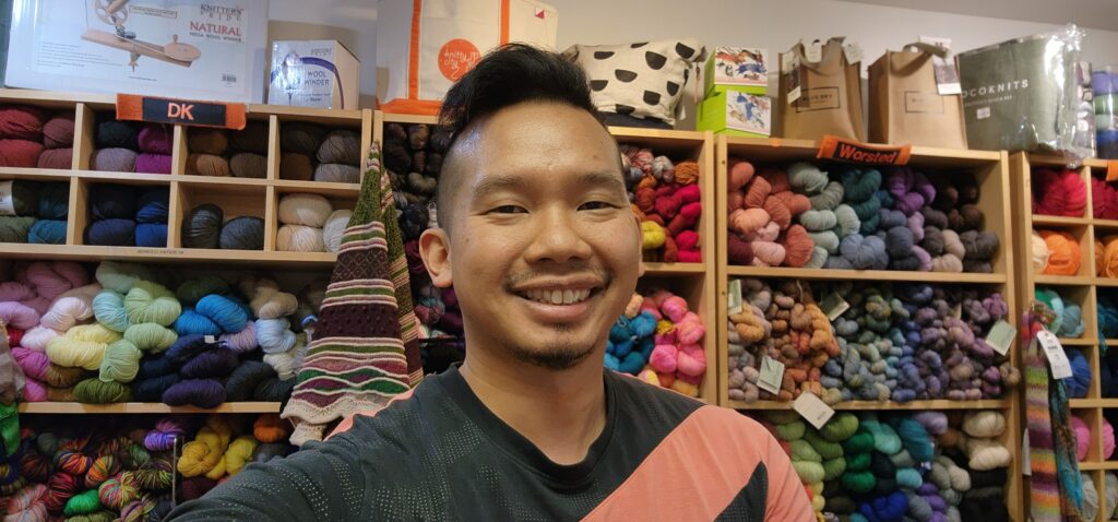 An Asian American man smiles in front of shelves of colorful yarn.