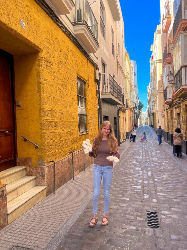 An alley between yellow buildings, and a light-skinned woman standing on the corner with blank yarn in her hands.
