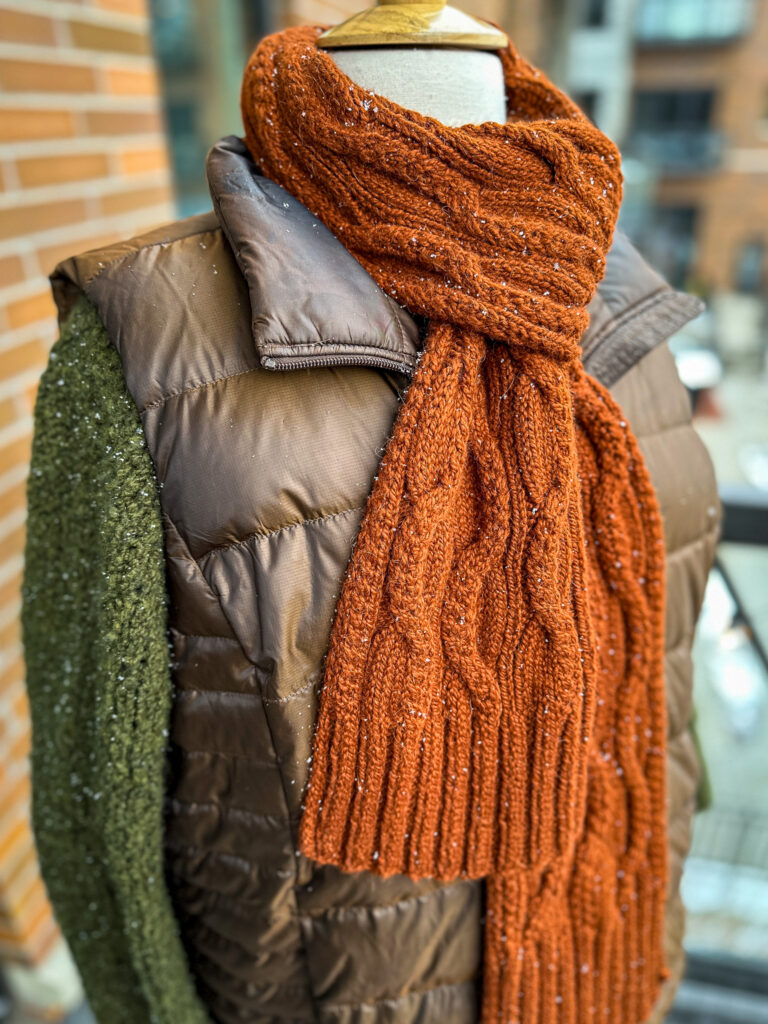 Rust orange cable scarf wrapped around dress form in front of brick wall.