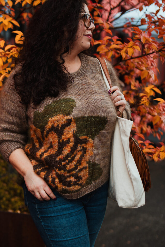 A light-skinned woman wearing a taupe sweater with an orange flower.