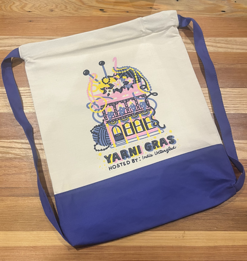A cream and royal blue backpack featuring an illustration of a New Orleans home decorated with yarn and beads and the text Yarni Gras, hosted by Indie Untangled.