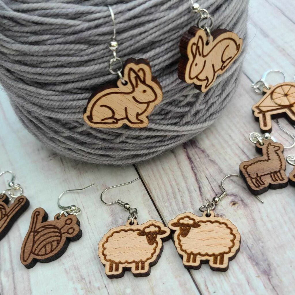 Wooden rabbit earrings on a cake of light grey yarn with knit/crochet, sheep, alpaca and spinning wheel earrings on the foreground.