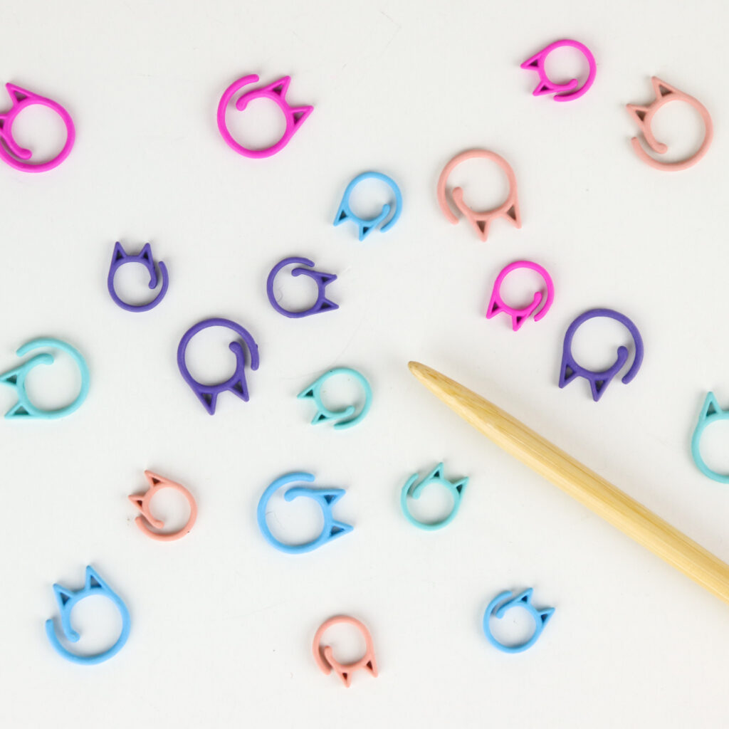 Colorful cat clips stitch markers spread out on a white background with a wooden knitting needle.