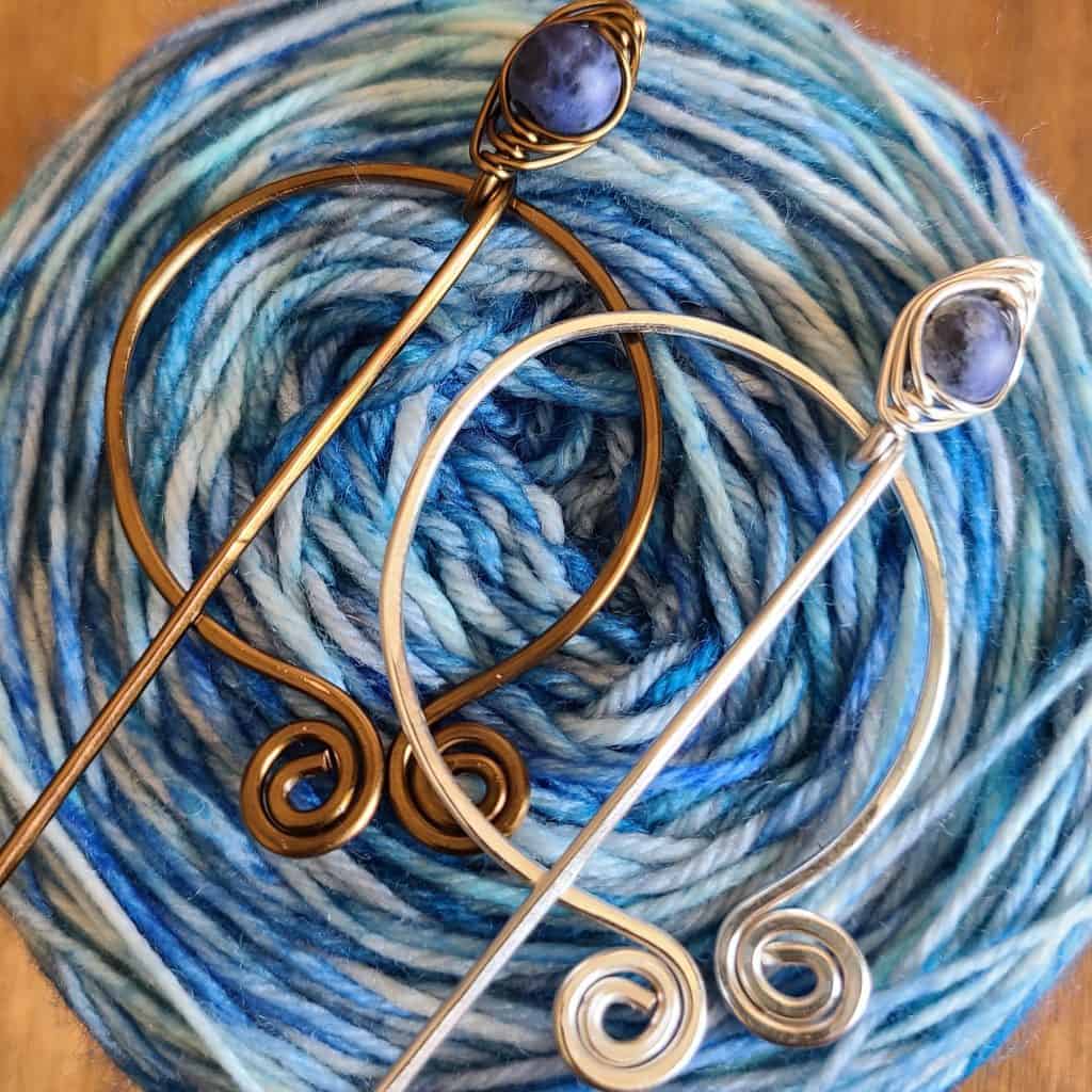 Shawl pins with blue stones in silver and bronze wire on a variegated blue yarn cake.