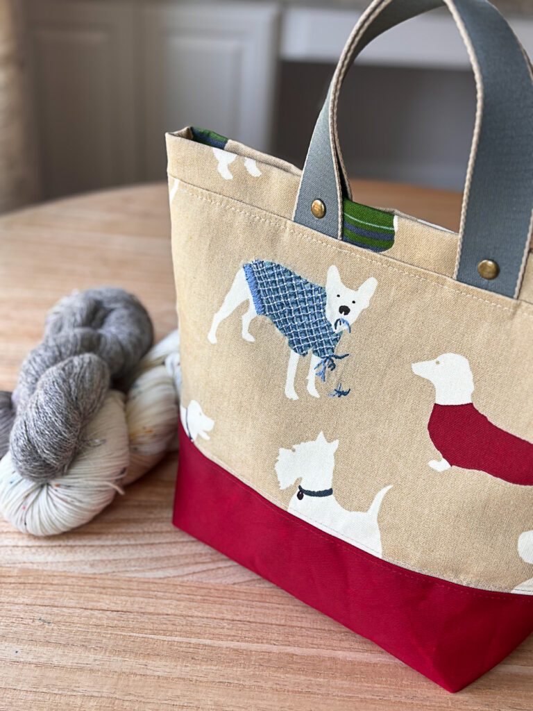 A bag with images of dogs on beige fabric and hanks of yarn on a wooden table.