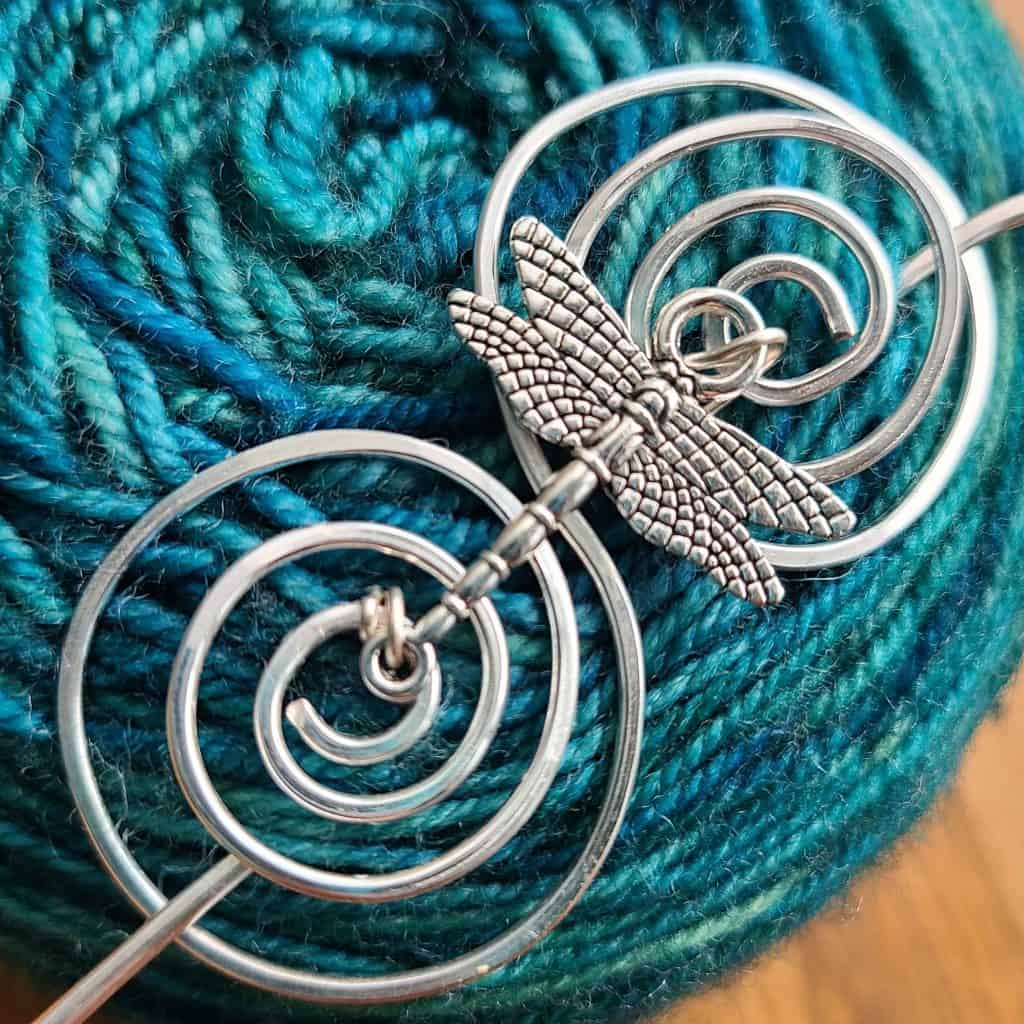 A silver dragonfly shawl pin on a forest green cake of yarn.