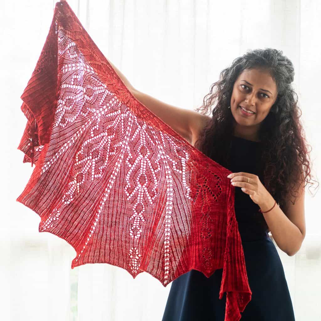 A brown-skinned woman holding up a large red knitted lace shawl.