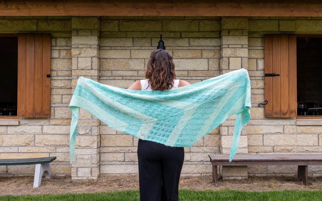 A young white woman holding white and blue shawl open across her back in front of a stone cottage.