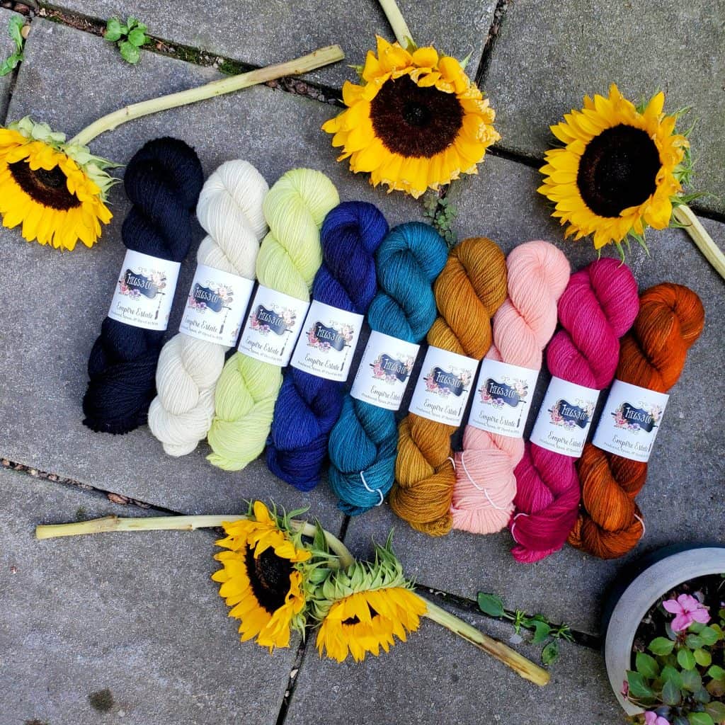 Skeins of yarn in assorted colors assembled in a row laying on a stone surface. Yellow sunflowers have been spread around them.