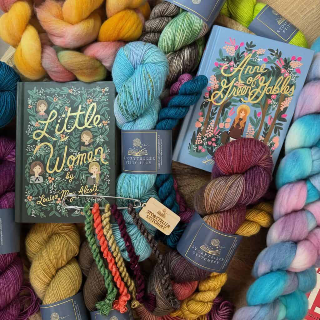 A flatlay showcasing hand-dyed yarn, fiber braids, and embroidery threads from the Cozy Hearthside Stories Collection, alongside Little Women and Anne of Green Gables books.