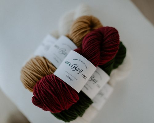 A stack of North Bay Fiber Heritage yarn in shades of red, brown, green and cream.