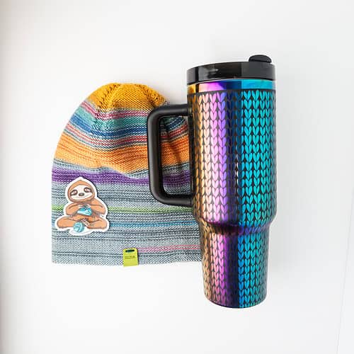 A knit hat featuring a soft, sewn-on microsuede tag that says "mine", a sticker of a sloth knitting, and a large tumbler that is engraved with a knit stitch pattern all over, revealing an iridescent rainbow metal underneath the black powder coating.