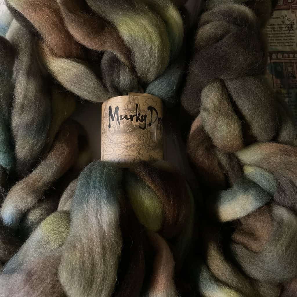 A bundle of fiber dyed in earthy, muted tones, such as olive green, brown, and soft blues. The label in the middle reads Murky Depths.