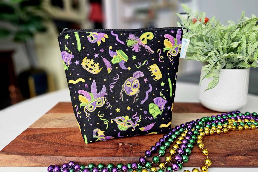 A black project bag with a vibrant Mardi Gras design featuring masks, crowns and beads, displayed alongside purple, green and gold bead necklaces.