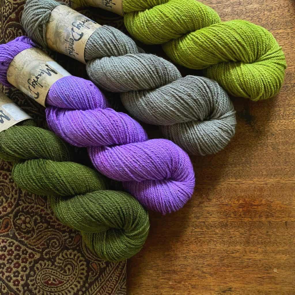 Skeins of pea green, mushroom, orchid and deep moss green yarn on a wooden table.