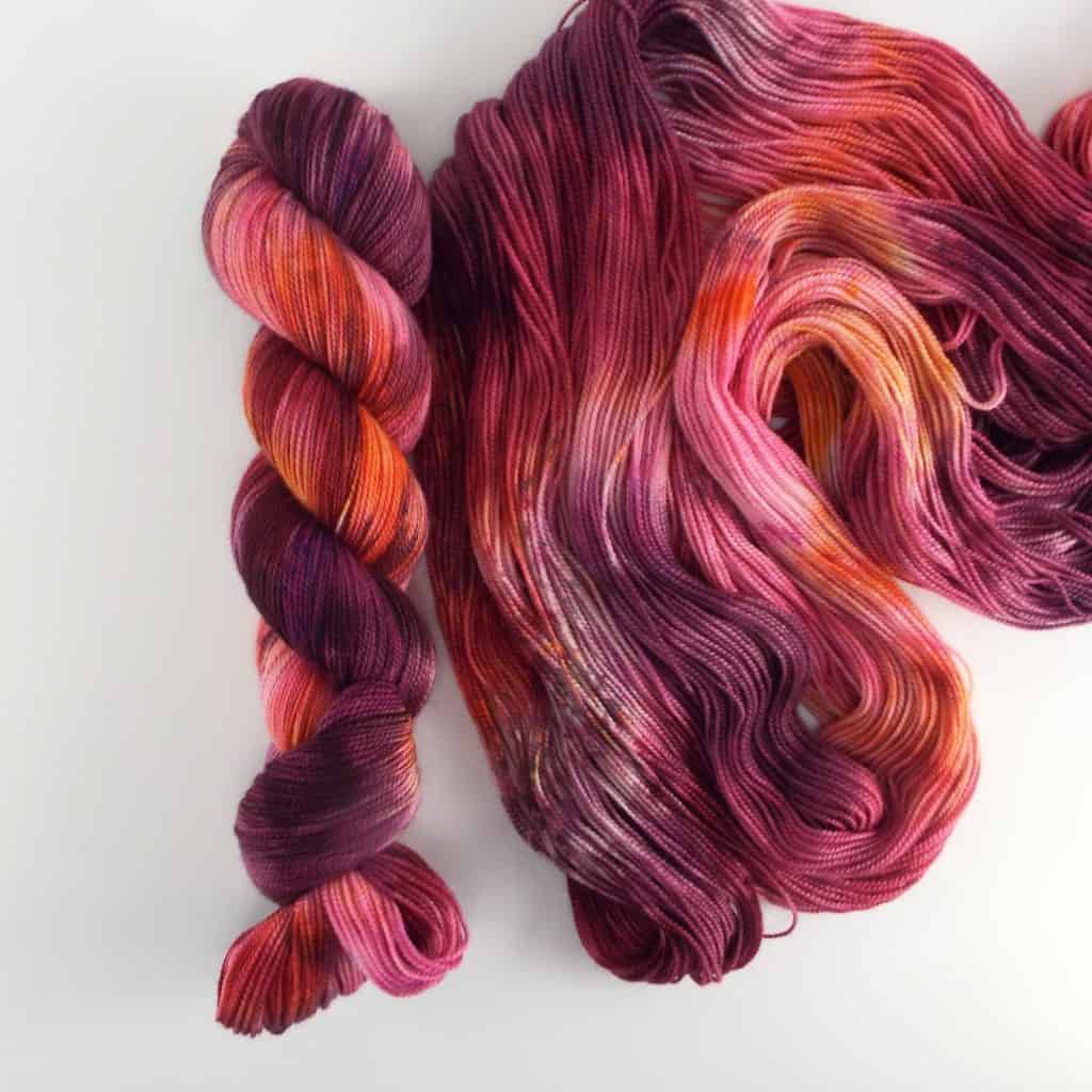 A single skein of yarn next to its unraveled strands. The yarn is hand-dyed with vibrant shades of magenta, plum, orange and pink, with subtle tonal variations.