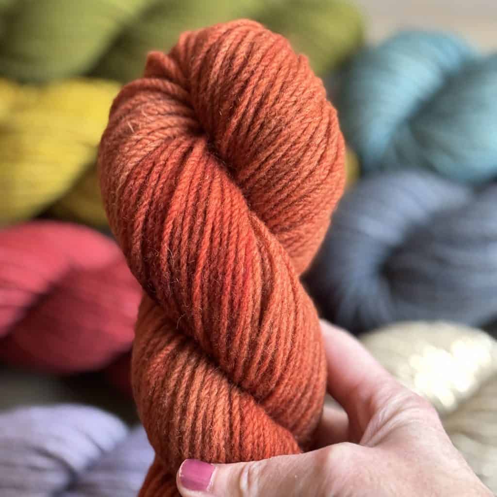 A close-up of an orange skein of yarn being held by a light-skinned hand, with various other colored skeins in the blurred background, including shades of yellow, green, blue, gray, and red.