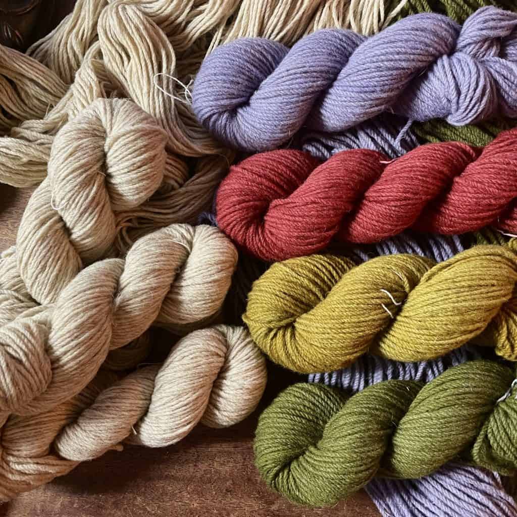 A collection of yarn skeins arranged on a wooden surface. The colors include natural cream with red undertones, soft lavender, deep red, mustard yellow, and olive green. Loose yarn strands in a neutral cream shade are spread in the background.