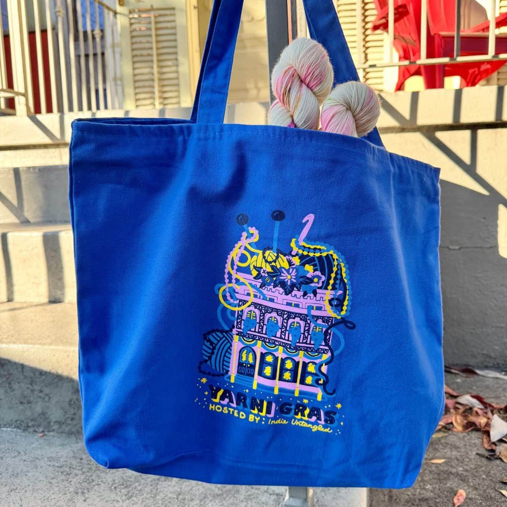 The Yarni Gras tote bag hanging on a metal post in front of a classic New Orleans-style home with green shutters and a red chair. The deep blue tote features an illustration of a New Orleans home decorated with yarn and beads in pink, navy, blue and yellow and the text Yarni Gras, hosted by Indie Untangled. Two skeins of hand-dyed yarn peek out from the top.