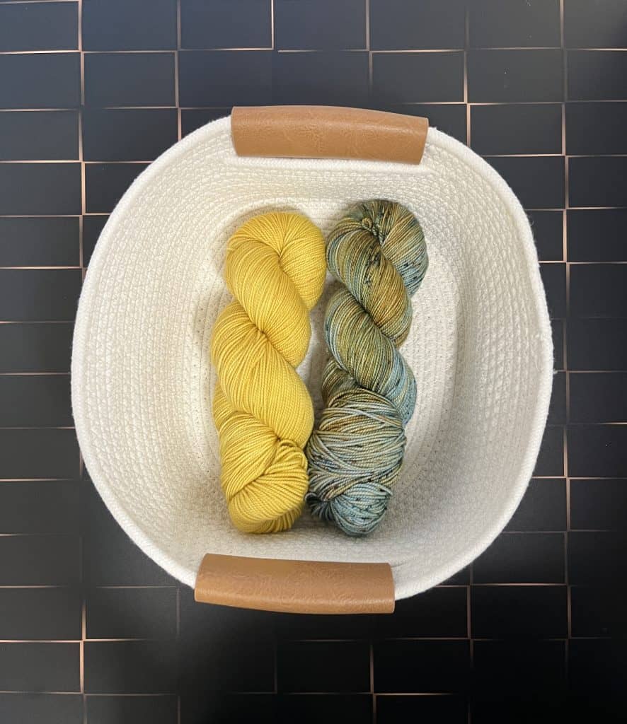 Two skeins of yarn (one golden yellow and one blue and yellow variegated) in a white rope basket on a black and gold background.