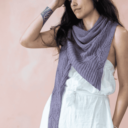 Woman wearing a hand-knitted purple shawl with a large cabled texture, draped over her shoulders. The shawl features V-shaped stitch details. She is dressed in a white belted dress with a statement necklace.