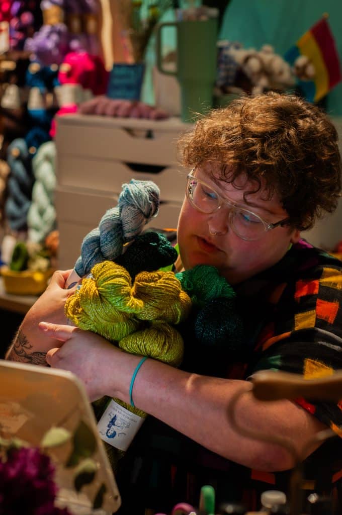 A light-skinned person with short curly brown hair and glasses holding several skeins of sparkly green and blue yarn close to their chest.