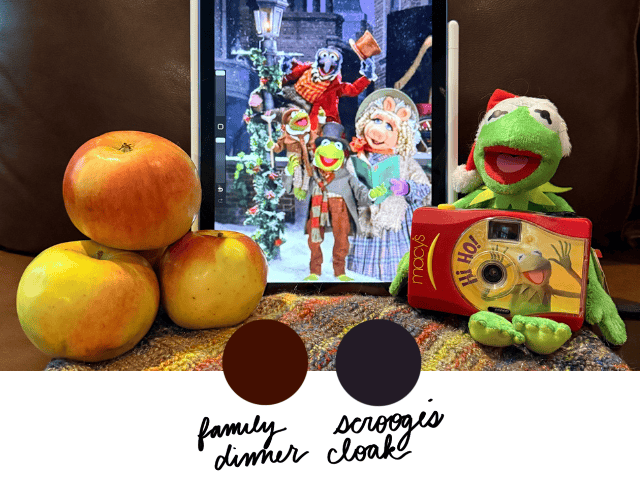 A display of apples, yarn, and a tablet showing characters from The Muppet Christmas Carol, with two color swatches labeled “Family Dinner” (deep brown) and “Scrooge’s Cloak” (dark navy).