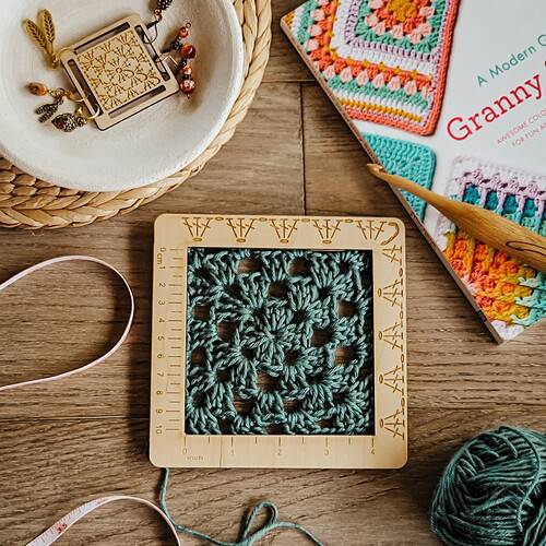 A teal granny square lays flat with a wooden gauge ruler on top with granny stitches engraved on it.