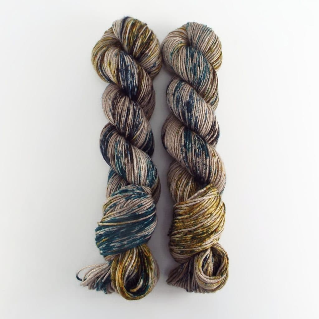 Two hand-dyed skeins in earthy beige tones speckled with teal, mustard, and dark brown, shown against a white background.