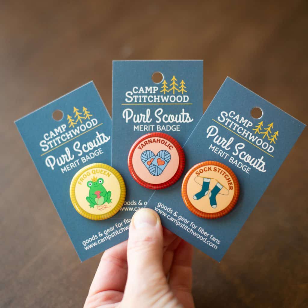 A hand holds up a fan of three buttons, each on a Camp Stitchwood backer