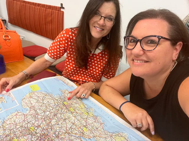 A woman with medium skin tone, dark hair, and glasses sits beside a woman with light skin tone, long brown hair, and glasses. They’re smiling while looking at a large paper map spread out on a table.