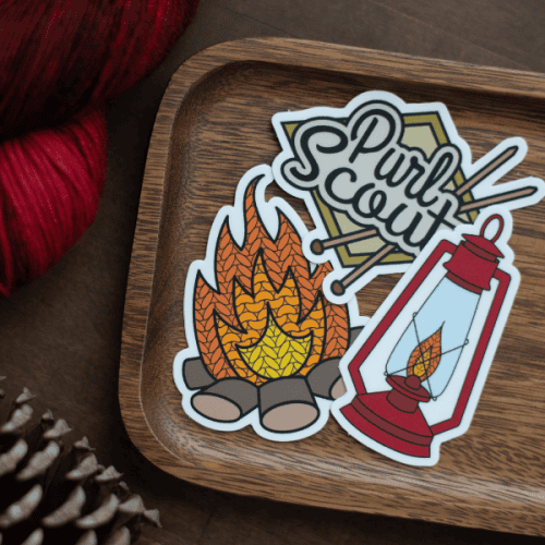 Three stickers of a lantern, a campfire, and the Purl Scouts logo sit on a wooden tray