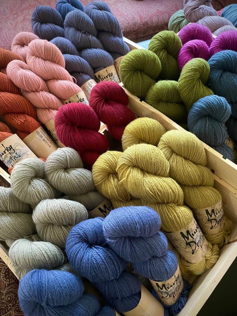 Skeins of yarn in rich jewel tones, including mustard yellow, sky blue, moss green, rose pink, red, and slate gray, arranged neatly in wooden crates on a patterned fabric background.