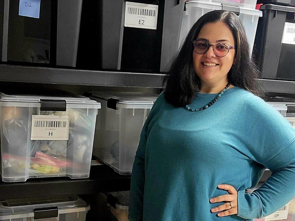 A woman with a light-medium skin tone and long dark hair, wearing a teal sweater and jeans, smiling with one hand on her hip while standing in front of shelves stacked with clear and black plastic bins of yarn.