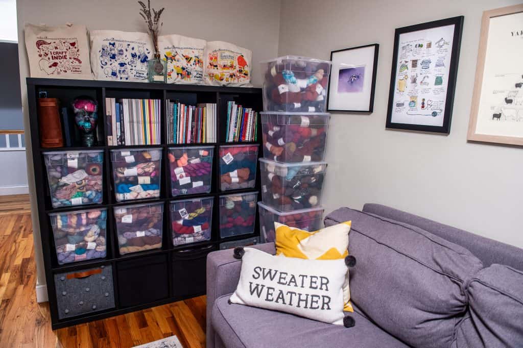 Black shelving units filled with clear bins of yarn skeins create a tidy studio backdrop, with framed fiber art on the wall and a gray sofa in the foreground.