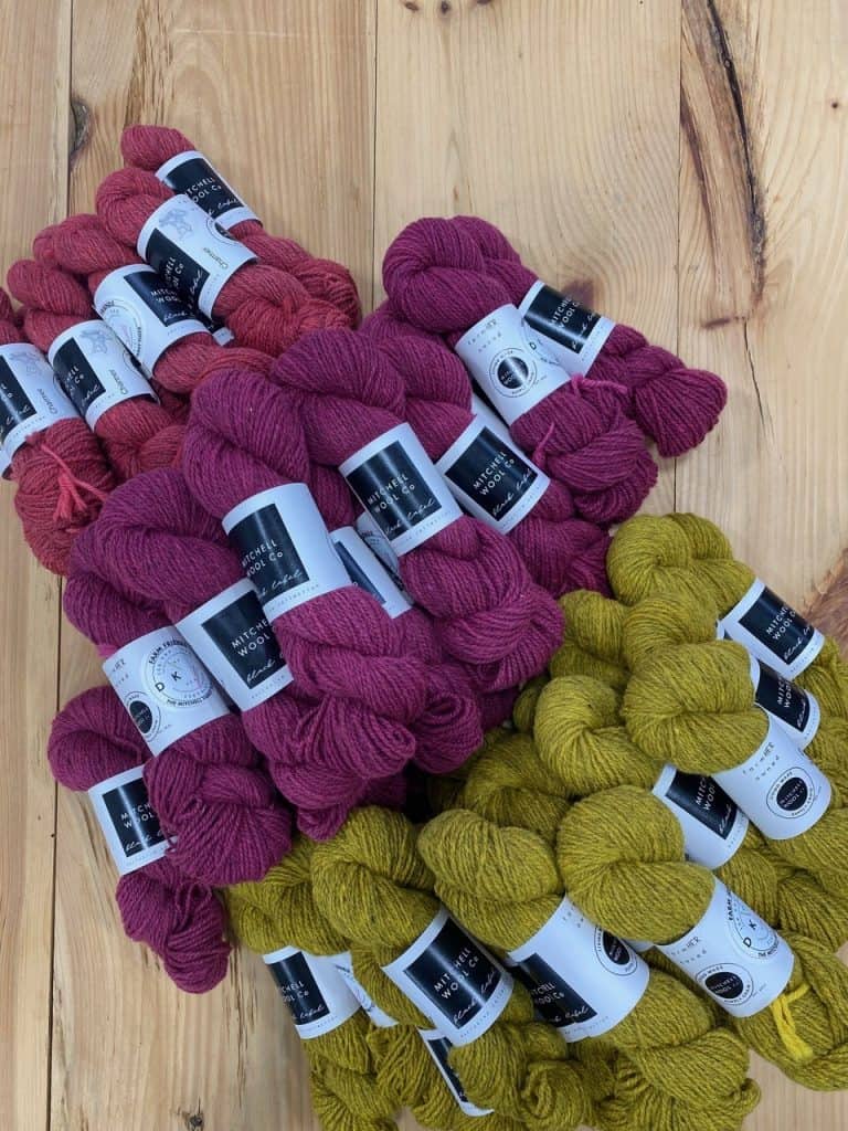 Stacks of magenta and olive green wool yarn skeins with branded labels, piled on a wooden tabletop in two color groups.