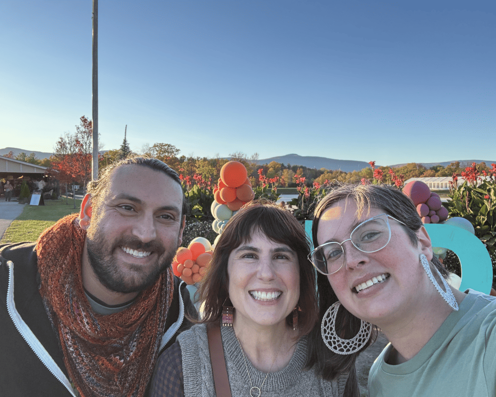 Three adults with light skin tones smile for a selfie outdoors at sunset, with colorful balloon decorations, flowers, and distant mountains behind them.