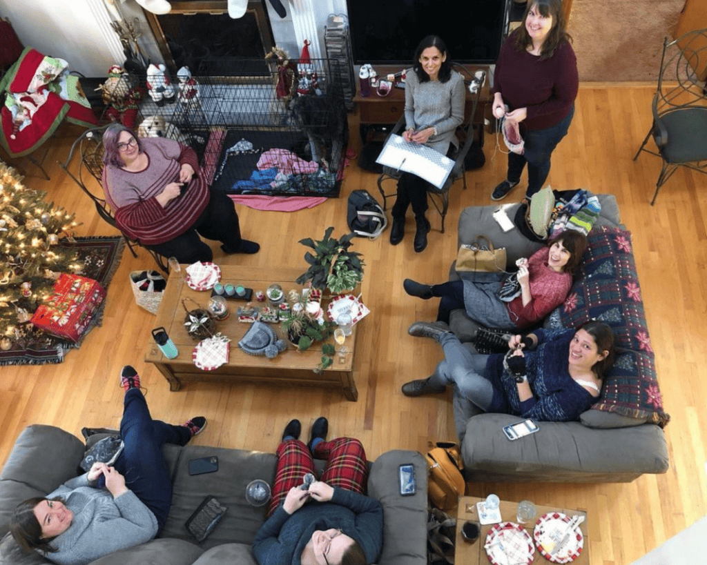 An overhead view shows a group of adults, mostly with light skin tones, gathered in a cozy living room knitting and chatting around a coffee table, with holiday decorations and a small dog in a crate nearby.