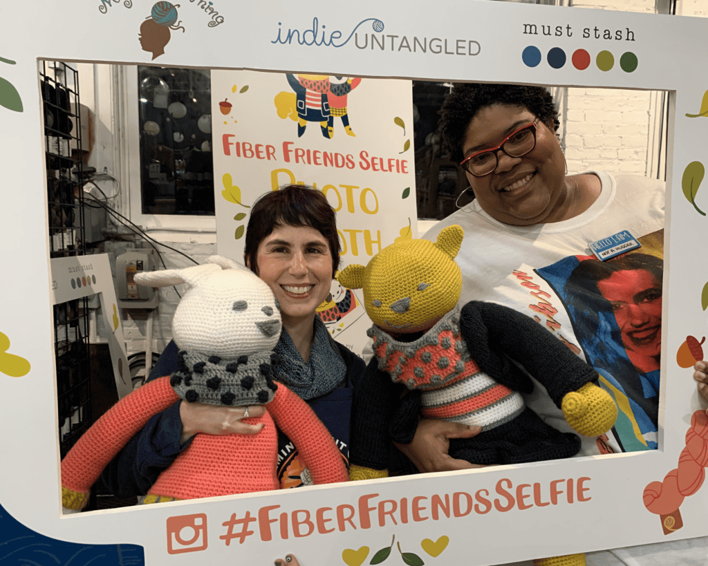 Two women pose indoors behind a “#FiberFriendsSelfie” frame; the woman on the left has a light skin tone, and the woman on the right has a dark skin tone. They smile while holding large knitted stuffed animals in a yarn shop setting.