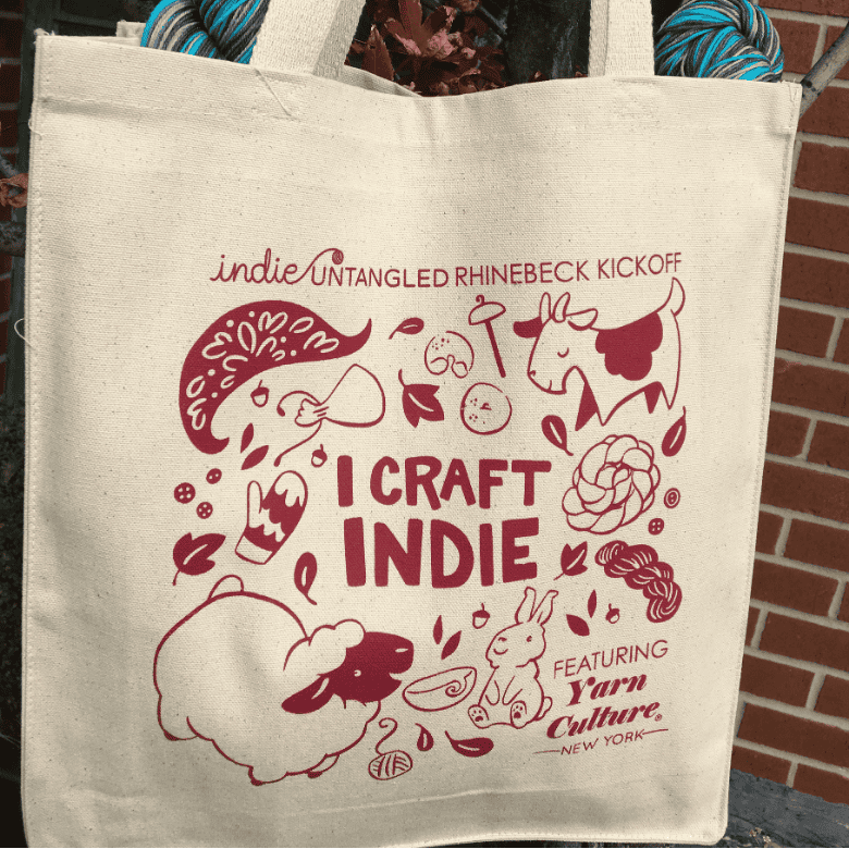 A cream-colored tote bag hangs outdoors against a brick wall, printed in red with the text “I Craft Indie” and illustrated with playful icons like a sheep, yarn, leaves, and crafting tools.