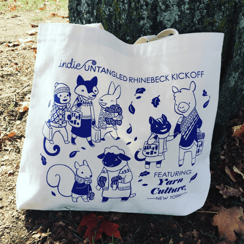 A cream tote bag rests against a tree outdoors, decorated in blue with cartoon animals wearing sweaters and carrying yarn, along with text referencing “Indie Untangled Rhinebeck Kickoff.”