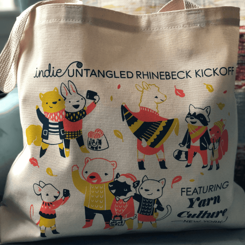 A cream tote bag displayed indoors features colorful illustrations of animals in scarves and sweaters, with text about the Indie Untangled Rhinebeck event and Yarn Culture New York.
