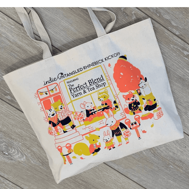 A cream tote bag lies flat on a wooden surface, showing a bright illustration of animals gathered around a yarn and tea shop, with text reading “The Perfect Blend Yarn & Tea Shop.”