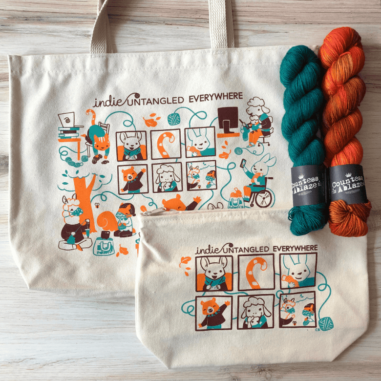 Two matching cream bags and two skeins of yarn are arranged on a wooden surface; the bags feature whimsical illustrations of animals crafting and using yarn, with the phrase “Indie Untangled Everywhere.”