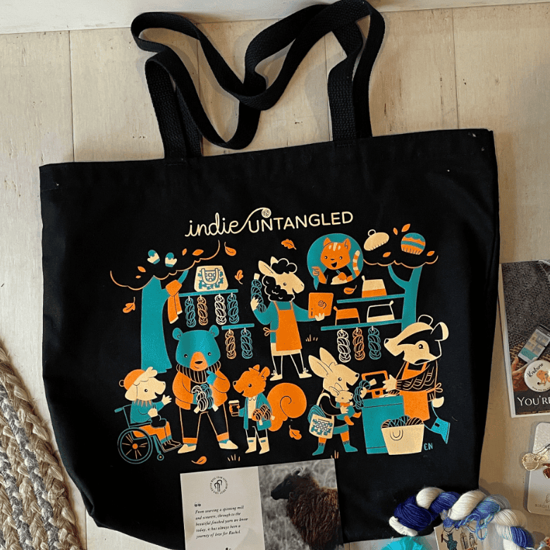 A large black tote bag is displayed indoors, featuring colorful illustrations of animals knitting and shopping for yarn, surrounded by small crafting items placed nearby.