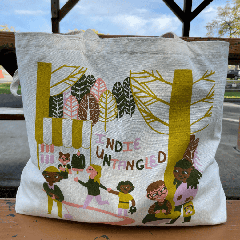 A cream tote bag sits on a bench outdoors, printed with a stylized forest scene and diverse illustrated people walking and shopping beneath trees, with “Indie Untangled” text in the center.