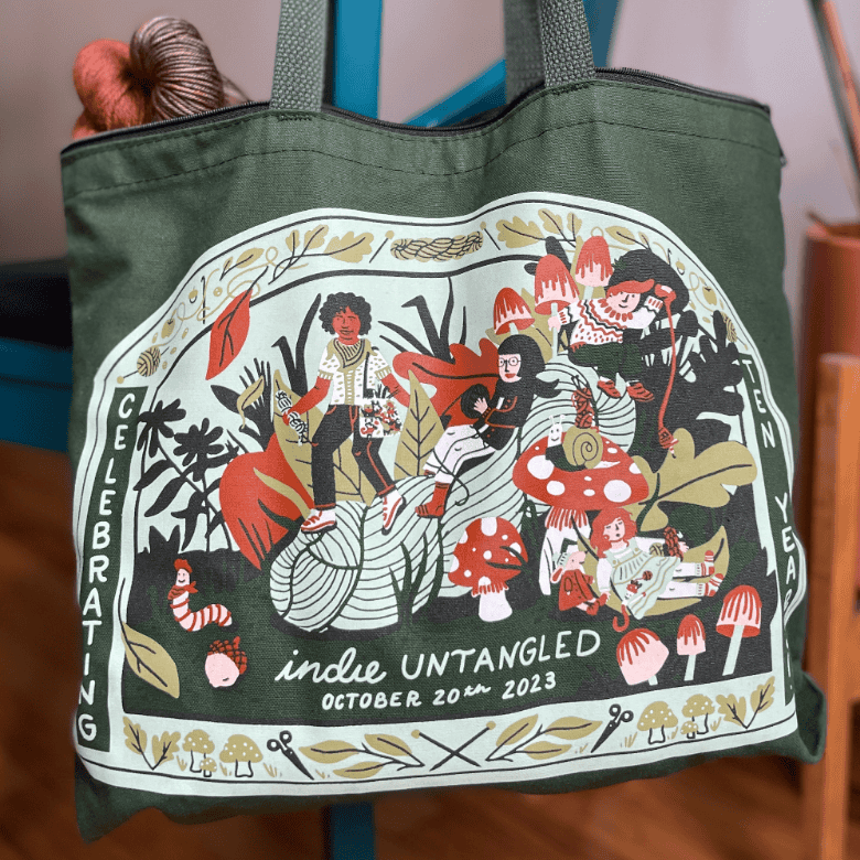 A green tote bag indoors features a detailed illustration of people crafting and relaxing among oversized yarn and mushrooms, with text reading “Indie Untangled October 20th 2023.”