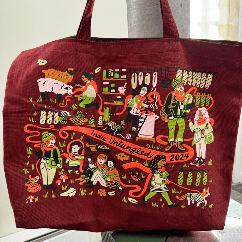 A red tote bag is shown indoors with a lively illustration of people and animals engaged in fiber arts, surrounded by yarn and tools, with “Indie Untangled 2024” across the design.