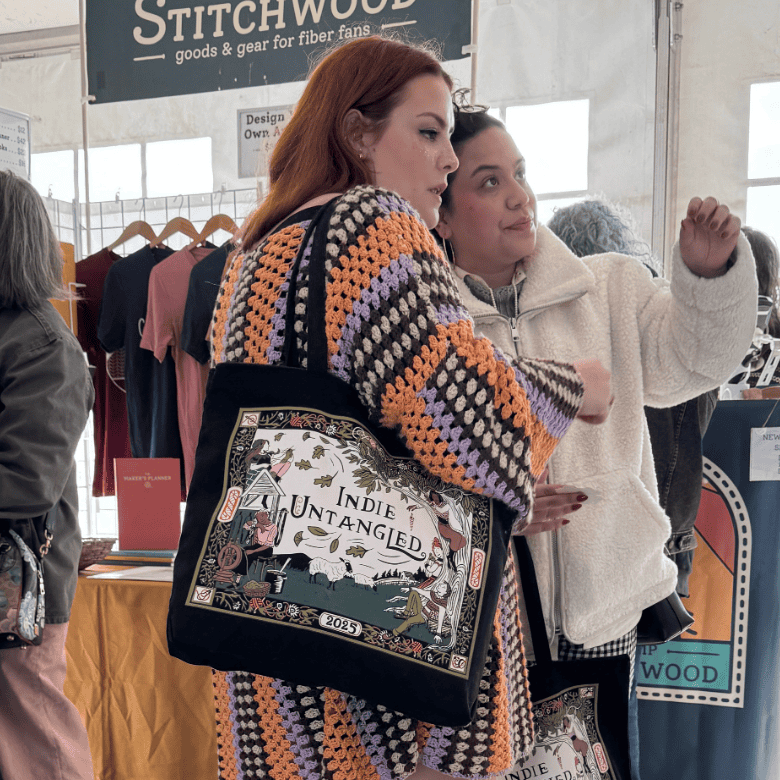 At a market booth, two people—one with a light skin tone wearing a colorful crocheted sweater and one with a medium skin tone in a light jacket—stand close together and point while looking at items for sale, both carrying illustrated Indie Untangled tote bags.